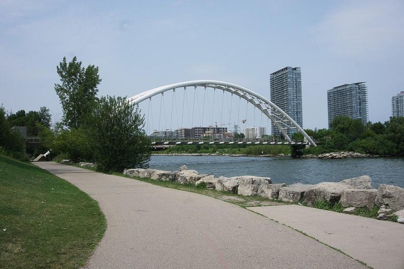 The Humber Bay Arch Bridge over the Humber River in Toronto with a paved waterfront path and condo towers beyond